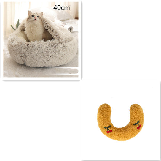 Cat sitting in a plush, round pet bed with attached blanket, alongside a yellow plush U-shaped neck pillow with embroidered cherries.